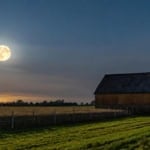 March Worm Moon astrology report represented by a full Moon beside a barn in early spring and green grass in the foreground.